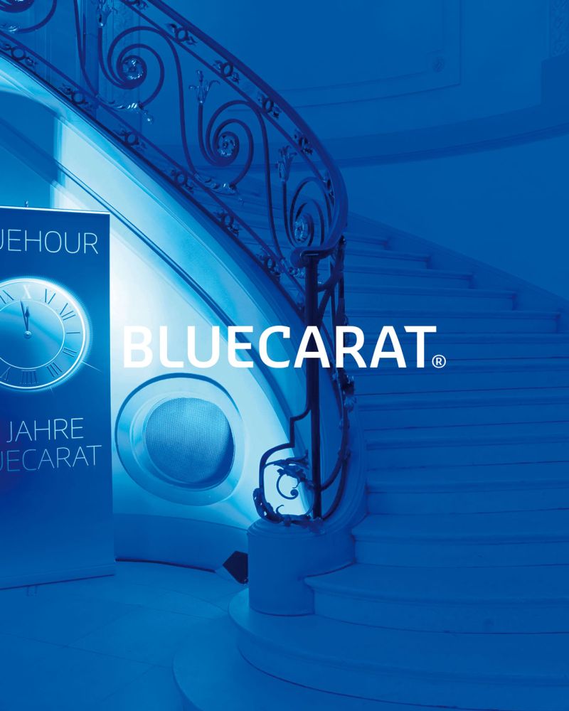 Bluecarat Logo vor blauem Hintergrund mit Rollup und Treppe auf einer Veranstaltung Bluecarat Logo vor blauem Hintergrund mit Rollup und Treppe auf einer Veranstaltung
