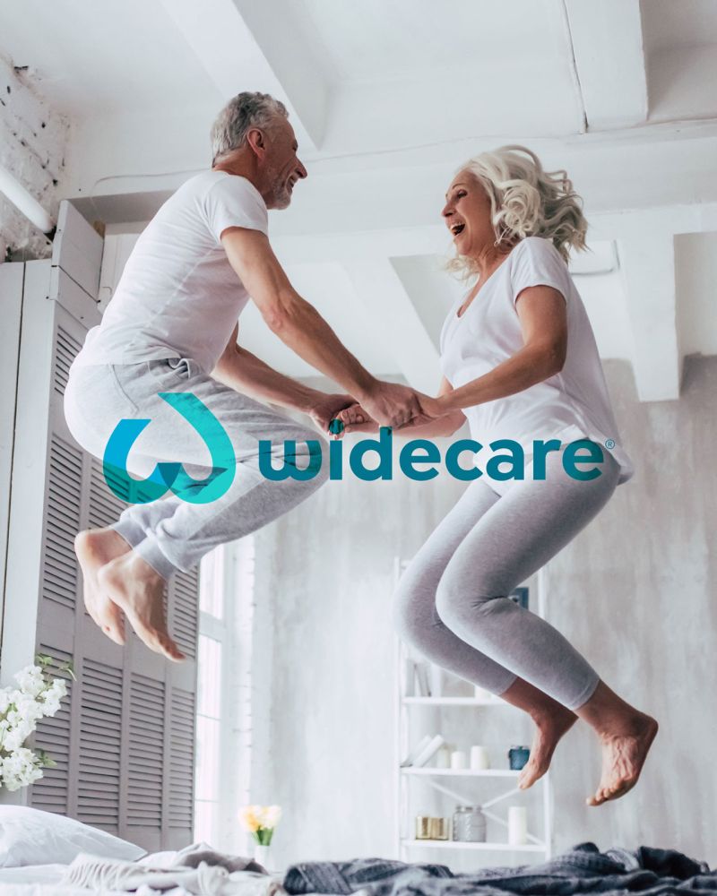 widecare Logo, Stuttgart, älteres Paar hüpft fröhlich auf Bett in Loft