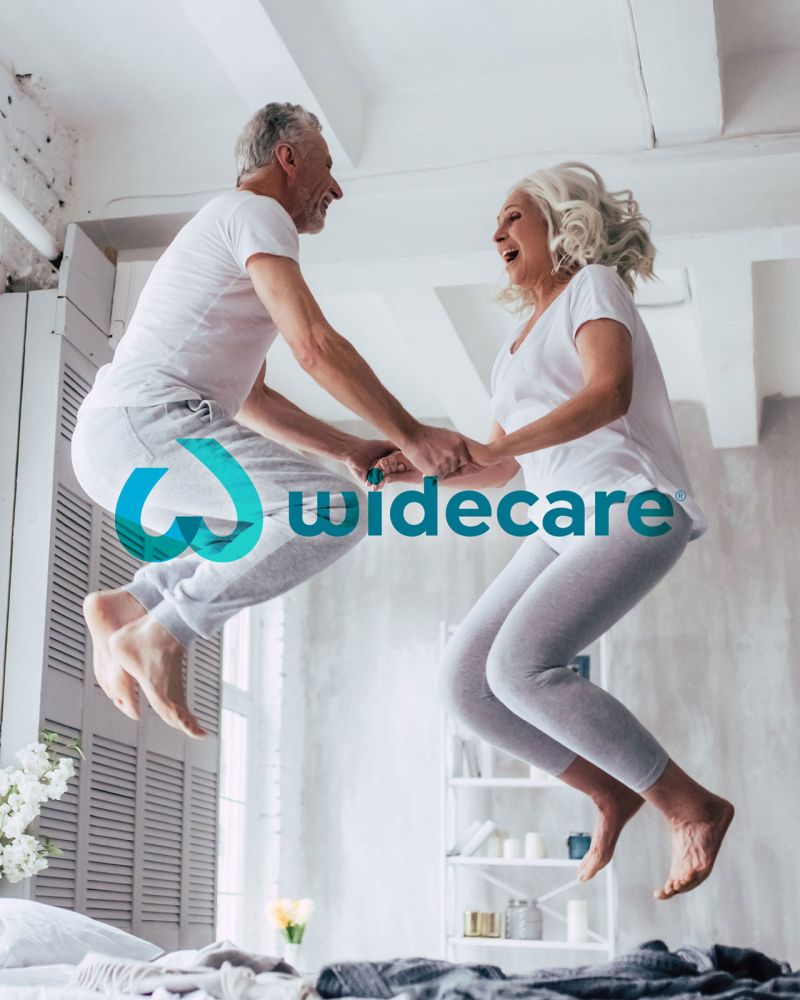 widecare Logo, Stuttgart, älteres Paar hüpft fröhlich auf Bett in Loft widecare Logo, Stuttgart, älteres Paar hüpft fröhlich auf Bett in Loft