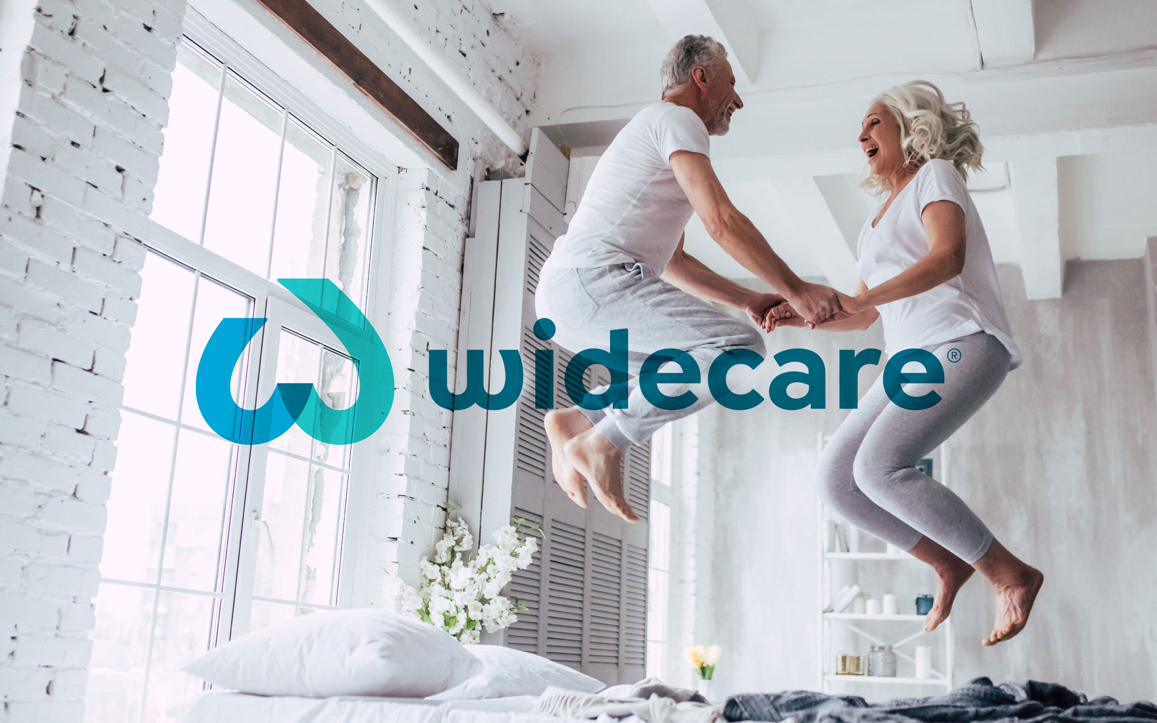 widecare Logo, Stuttgart, älteres Paar hüpft fröhlich auf Bett in Loft widecare Logo, Stuttgart, älteres Paar hüpft fröhlich auf Bett in Loft